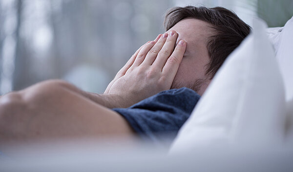 Schlafapnoe geplagter Mann liegt im Bett und hält sich die Hände vors Gesicht