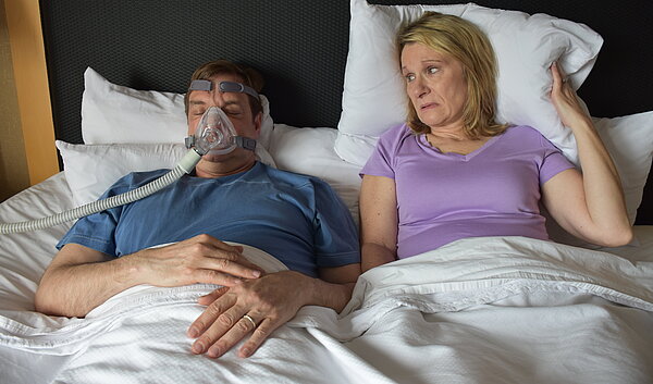 Ehepaar liegt im Bett, Partner trägt eine CPAP Maske