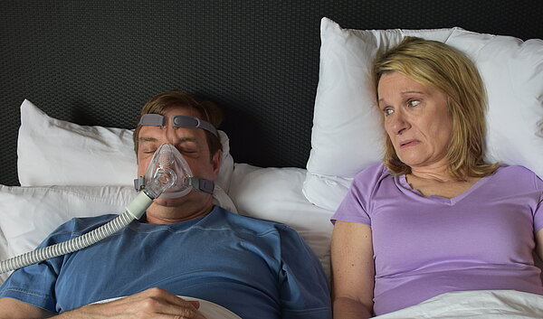 Ehepaar im Bett, Partner trägt eine CPAP Maske.
