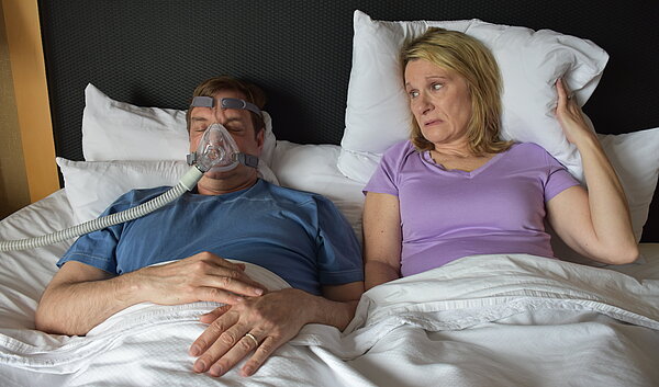 Ehepaar liegt im Bett, Partner trägt CPAP Maske